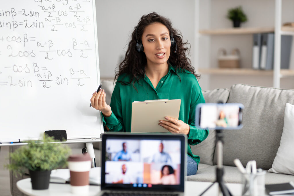 A female-presenting teacher in a green blouse with a headset on in a cozy setting with a whiteboard behind them and a clipboard in the hands talking to their cellphone on a small tripod on the table that is streaming them to a video call with students that you can see on an opened laptop blurred a bit in the foreground.