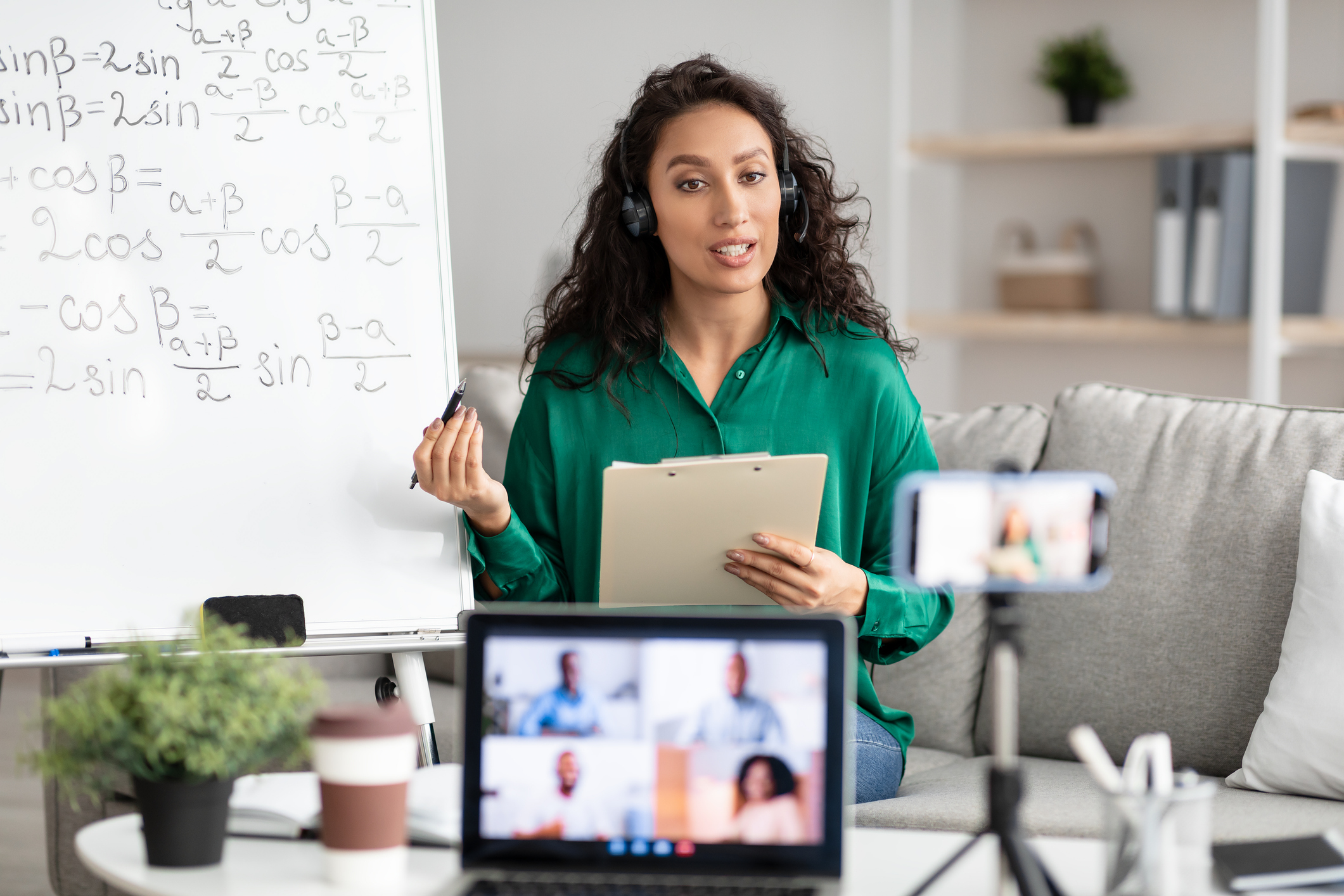 A female-presenting teacher in a green blouse with a headset on in a cozy setting with a whiteboard behind them and a clipboard in the hands talking to their cellphone on a small tripod on the table that is streaming them to a video call with students that you can see on an opened laptop blurred a bit in the foreground.