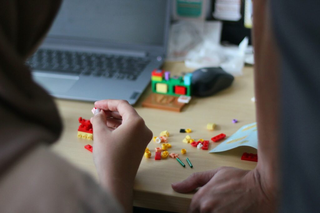 Hands playing with colourful LEGO block pieces on a wooden table with a laptop open in the background.
