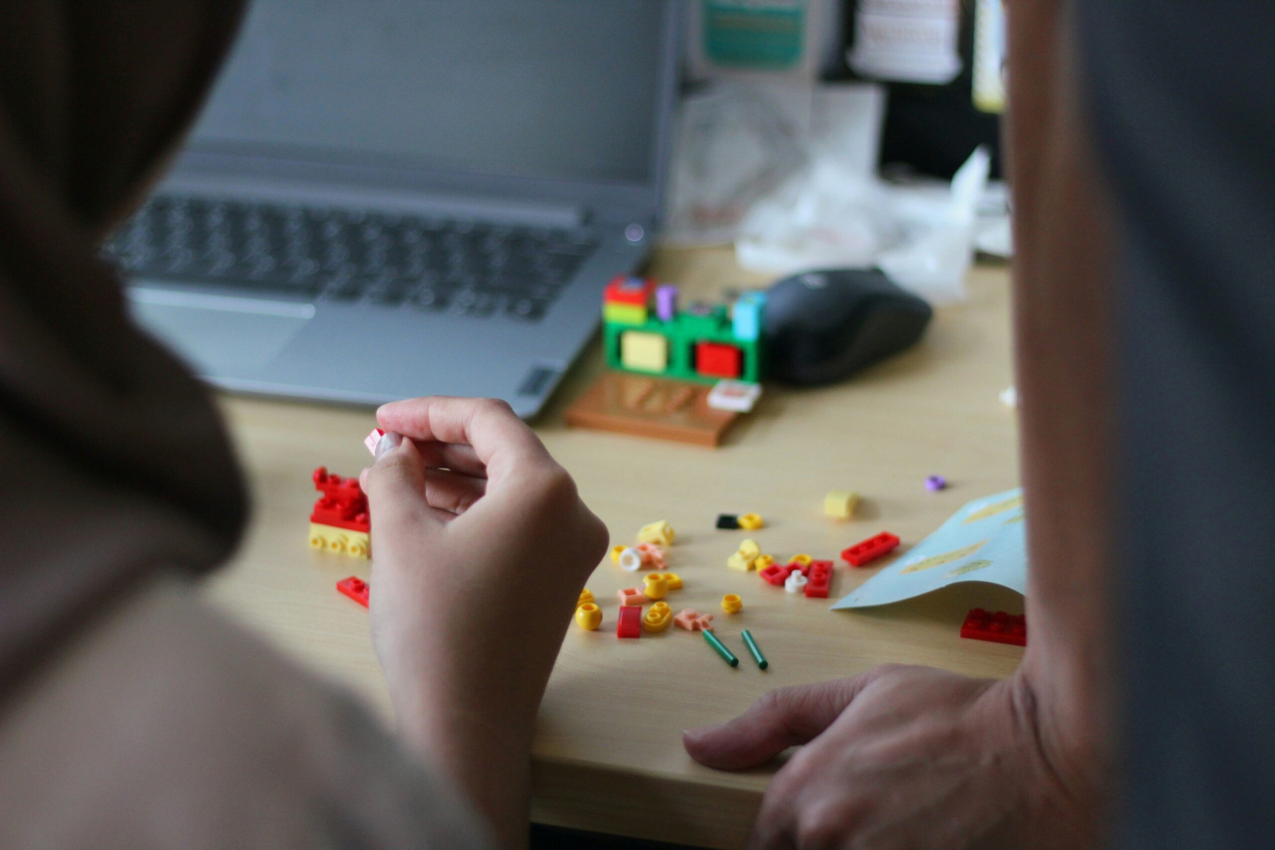 Hands playing with colourful LEGO block pieces on a wooden table with a laptop open in the background.